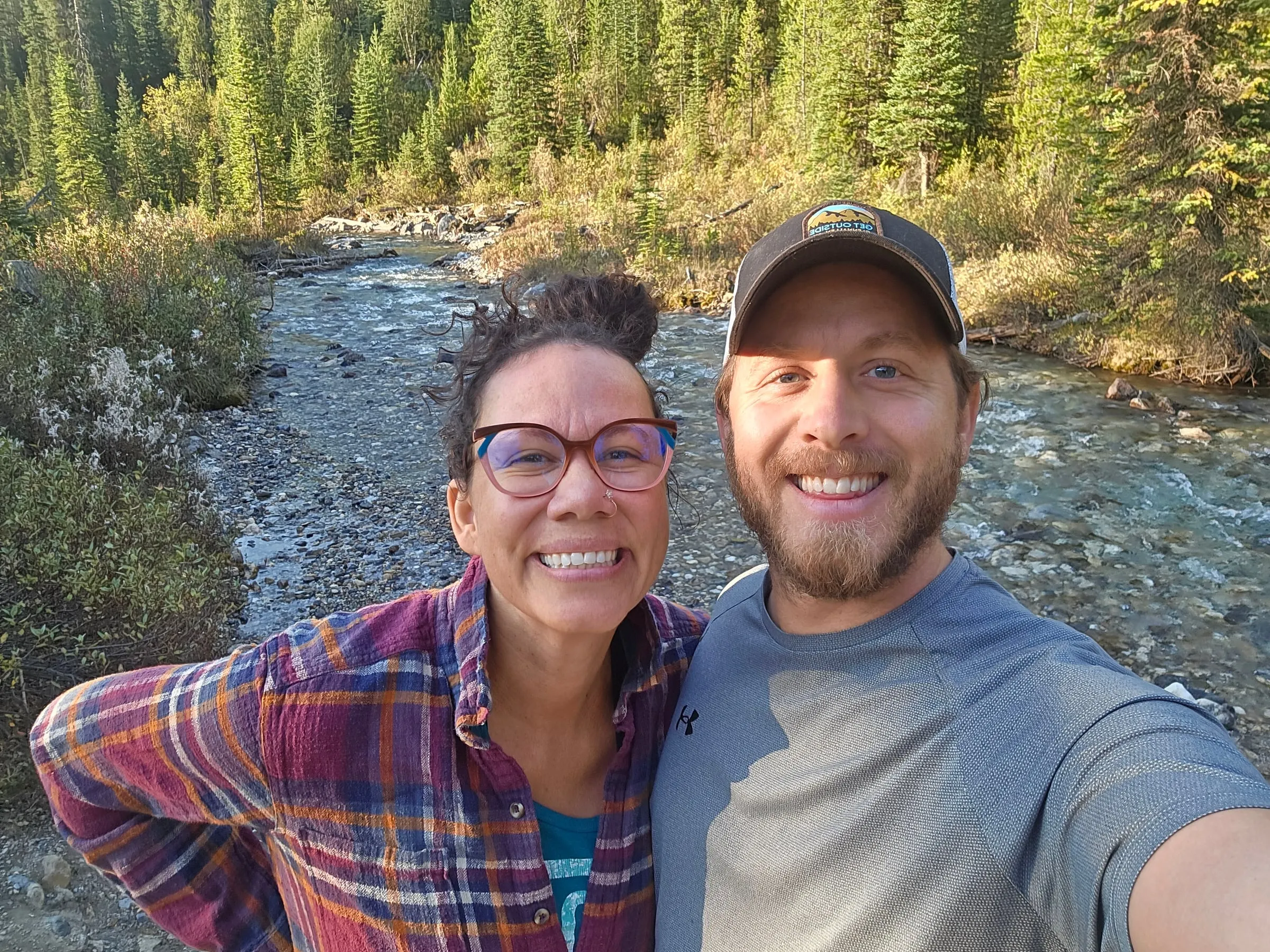 Two smiling people taking a selfie beside a river in the woods